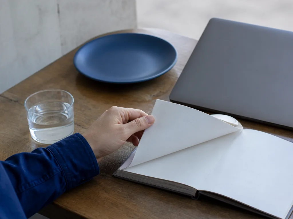 Photorealistic editorial photograph of a customer’s hand flipping through a blank paper notebook at a wooden café table,